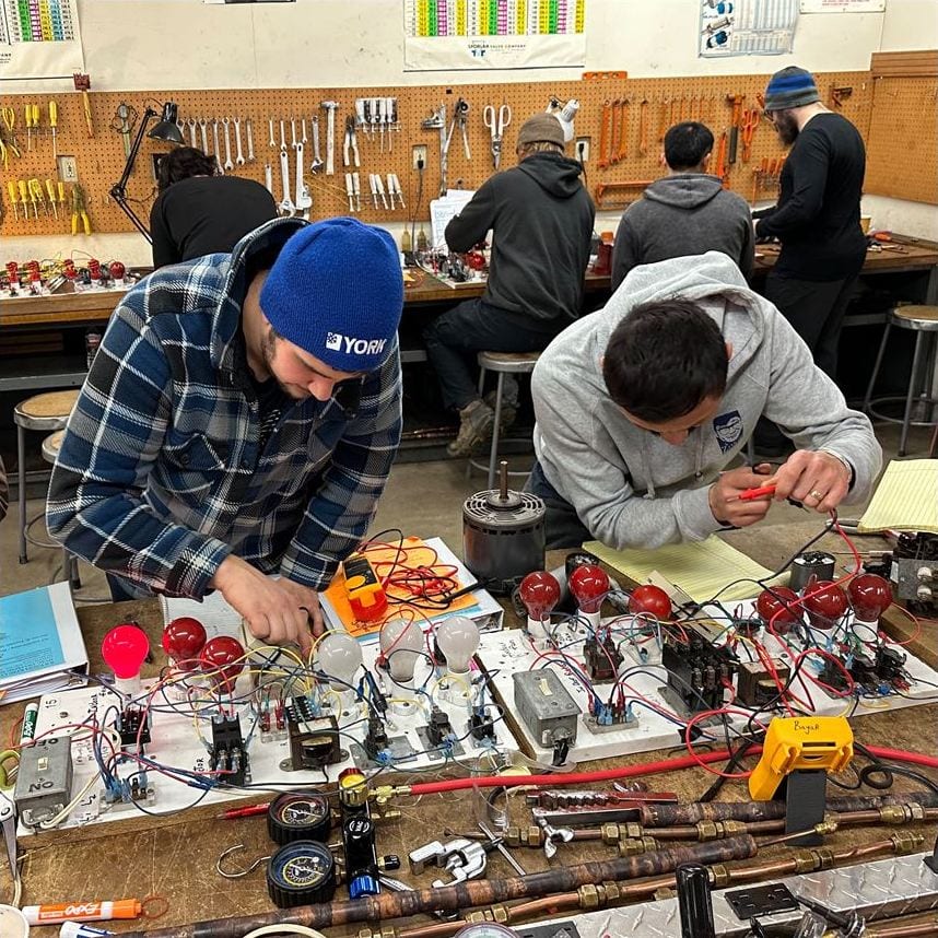 Two HVAC School students wiring components and reading meters at an electrical training bench